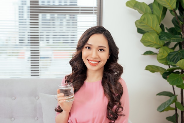 Молодая красивая азиатская женщина расслабляясь в софе с чашкой чистой воды