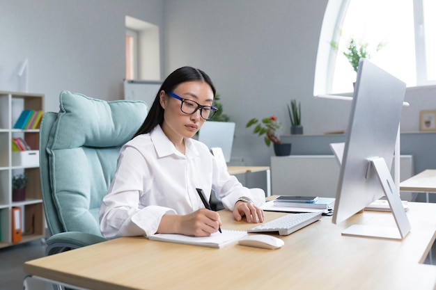 Young beautiful asian woman secretary making notes in notebook with pen sitting at desk in office