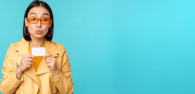 Young beautiful asian woman showing credit card smiling choosing bank standing over blue background