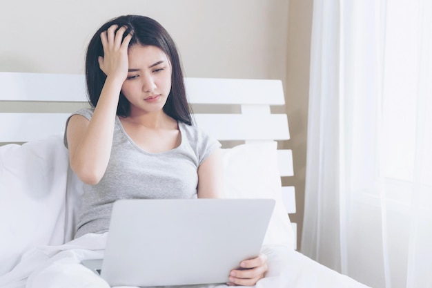 ベッドに座っている若い美しいアジアの女性彼女はラップトップを使用して深刻な感じと頭痛