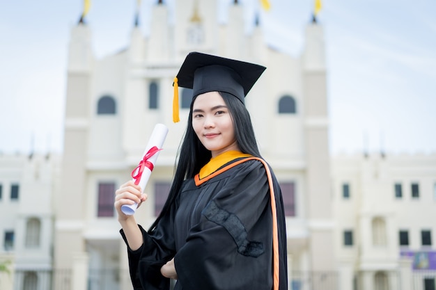 A young beautiful asian woman university graduate in graduation\
gown and mortarboard holds a degree certificate stands in front of\
the university building after participating in college\
commencement