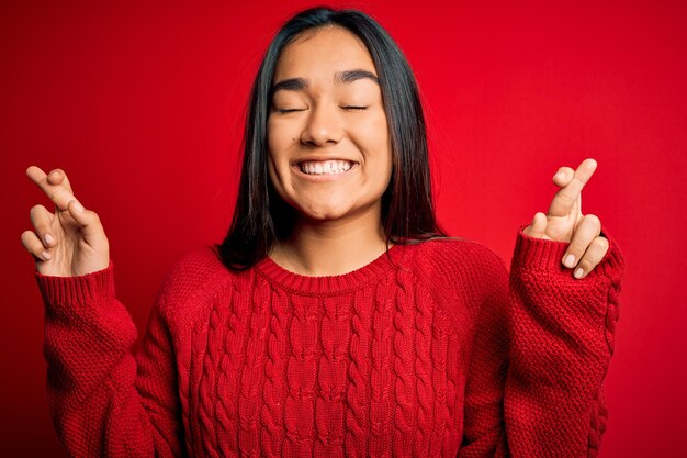 Young beautiful asian woman wearing casual sweater standing over isolated red background gesturing finger crossed smiling with hope and eyes closed luck and superstitious concept