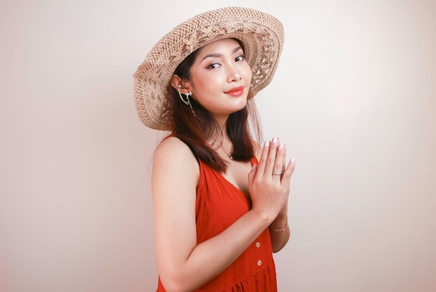 A young beautiful asian woman wearing a straw hat gives\
greeting hands with a big smile on her face indonesian woman on\
white background