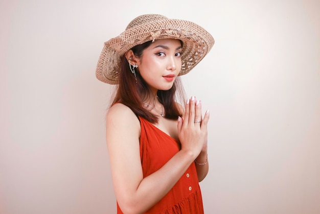 A young beautiful asian woman wearing a straw hat gives\
greeting hands with a big smile on her face indonesian woman on\
white background