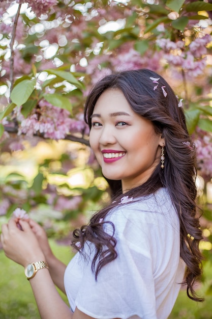 A young beautiful Asian woman in a white dress walks in a flowered park. Sakura. Blooming trees. Spring.