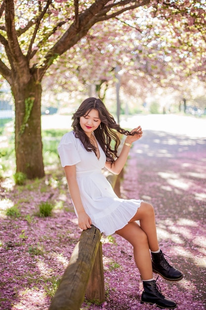 A young beautiful Asian woman in a white dress walks in a flowered park. Sakura. Blooming trees. Spring.