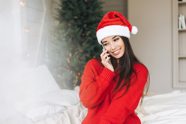 Young beautiful asian woman with dark long hair in cozy red
knitted sweater and santa hat using mobile smartphone on bed in
room with christmas tree at the home