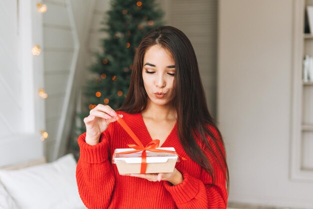 Young beautiful asian woman with dark long hair in cozy red\
knitted sweater and santa hat with present gift box with red ribbon\
sitting on bed in the room with christmas tree