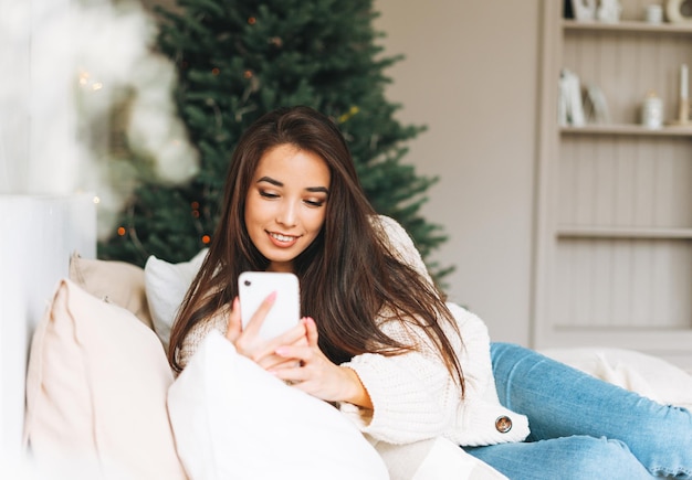 Young beautiful asian woman with dark long hair in cozy white knitted sweater using mobile smartphone on bed in the room with Christmas tree