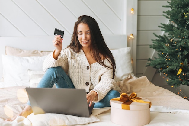 Young beautiful asian woman with dark long hair in cozy white\
knitted sweater with present gift box with golden ribbon using\
laptop with credit card on bed in room with christmas tree at\
home