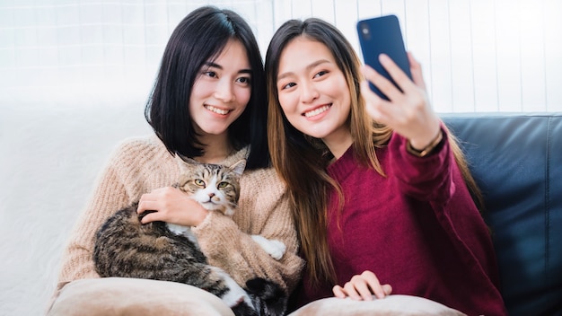 Young beautiful Asian women lesbian couple lover using smartphone selfie cute cat pet in living room at home with smiling face. Concept of LGBT sexuality with happy lifestyle together.