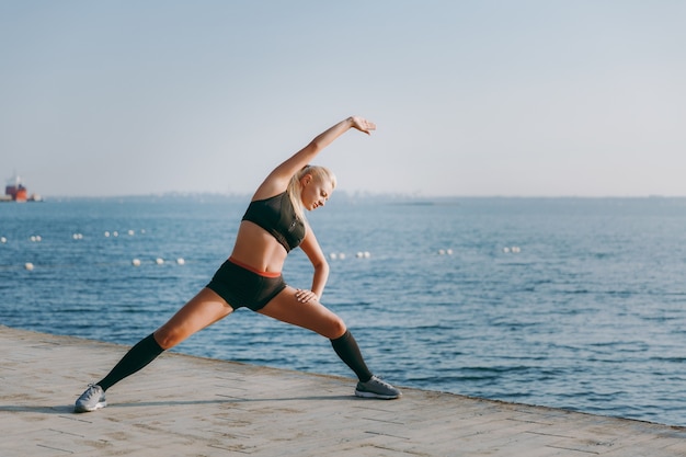 La giovane bella ragazza atletica con lunghi capelli biondi in abiti neri che si allena e fa esercizi, alzando il braccio all'alba sul mare