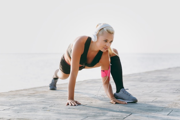 La giovane bella ragazza atletica con lunghi capelli biondi in cuffia e telefono cellulare in mano ascoltando musica e facendo stretching all'alba sul mare