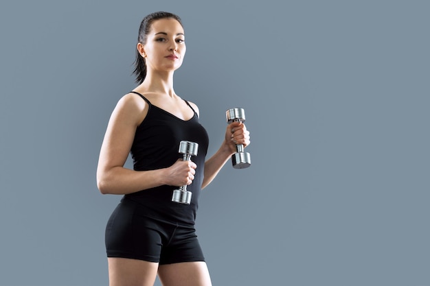 Young beautiful athletic muscular woman doing sports exercises using dumbbells, gray studio background, copy space