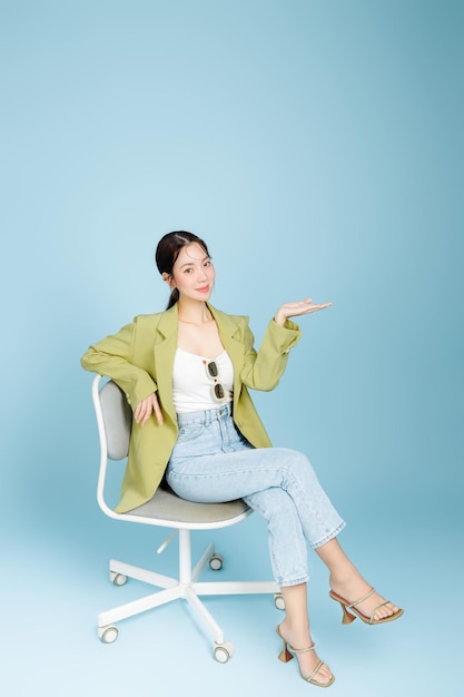 Photo young beautiful attractive asian woman in casual green fashion jacket and sunglasses sitting on armchair showing copy space isolated on pastel blue background female consumerism lifestyle concept