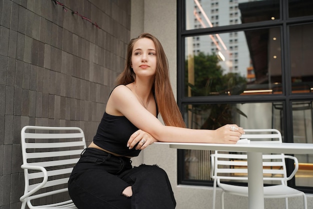 Young beautiful and attractive woman with long brown hair in a black dress at cafe against urban background