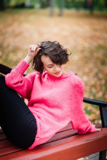 Young beautiful ballerina in fair coat and pointe shoes sit on the bench, rest outdoors in autumn park.