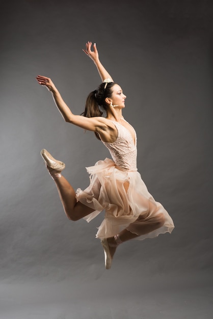 Young beautiful ballet dancer in beige swimsuit posing on pointes on light grey studio background