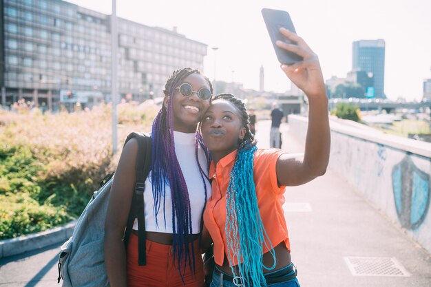 Young beautiful black sisters outdoor backlight using smartphone taking selfie