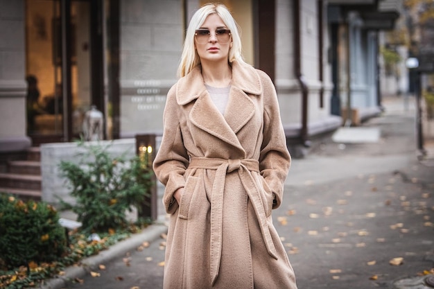 Photo young beautiful blond woman in an autumn coat stands on a city street. soft focus. fashionable concept. gray background