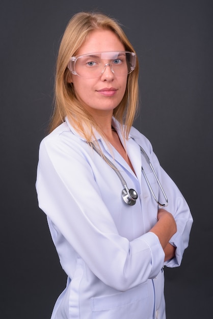  young beautiful blond woman doctor as scientist with protective glasses against gray wall