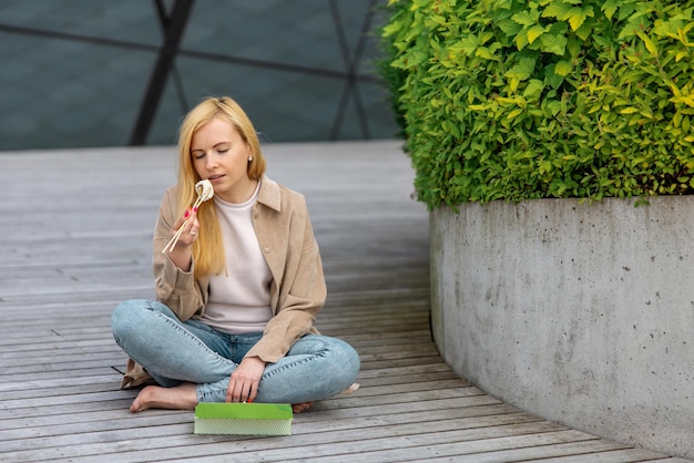 Young beautiful blond woman eating sushi outdoors on the wooden terrace by modern building in the city Tasty food to go Girl has lunch break spending time outside and eating Asian food City life