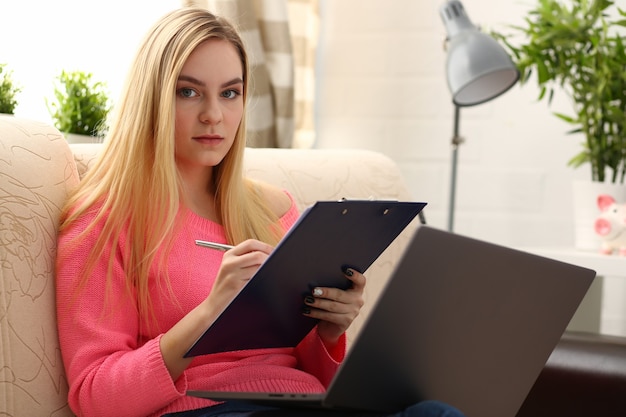 Young beautiful blond woman sit on the sofa in livingroom hold binder in arms work on laptop