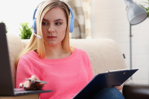 young beautiful blond woman sit on the sofa in livingroom hold binder in arms work with laptop listen to music