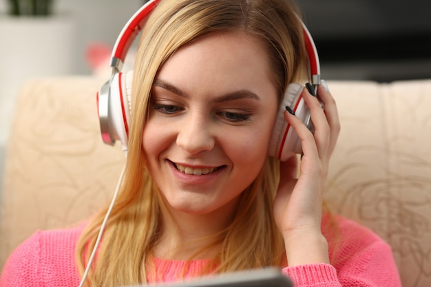 Young beautiful blond woman sit on the sofa in livingroom hold tablet in arms listen to music