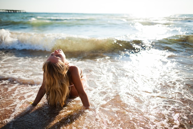 波の海の水の端に座っている白いビキニの若い美しいブロンドの女性