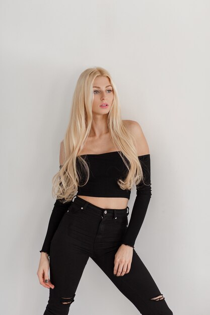 Young beautiful blond woman with freckles in black fashionable clothes stands near the white wall