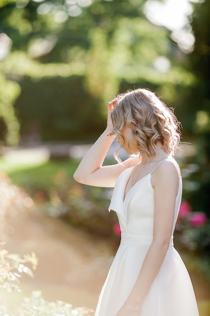 Молодая красивая блондинка 20 лет с вьющимися волосами в белом платье в солнечных лучах заката в парке Стильная модель гуляет по цветущему городскому парку летом