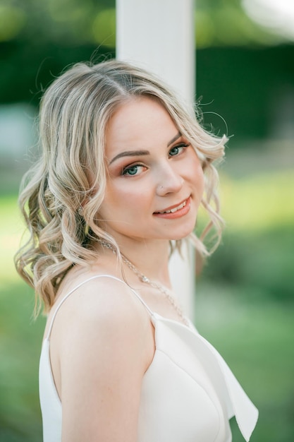 Giovane bella bionda anni '20 con i capelli ricci in un abito bianco nei raggi del sole del tramonto nel parco un modello elegante cammina attraverso il parco fiorito della città in estate