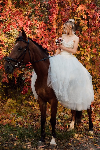가을 숲에서 승마를 덮고 있는 하얀 솜털 드레스를 입은 젊은 아름다운 금발 신부