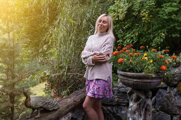 Young beautiful blonde girl in a beige knitted sweater and pink dress stands hugging herself