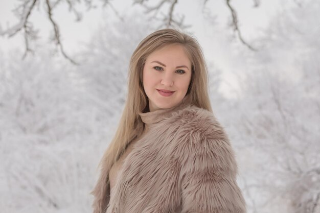 a young beautiful blonde girl in a fur coat stands in a snowy park
