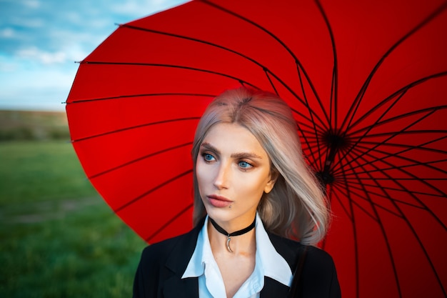 Young beautiful blonde girl, holding red umbrella, wearing black suit with choker on neck.