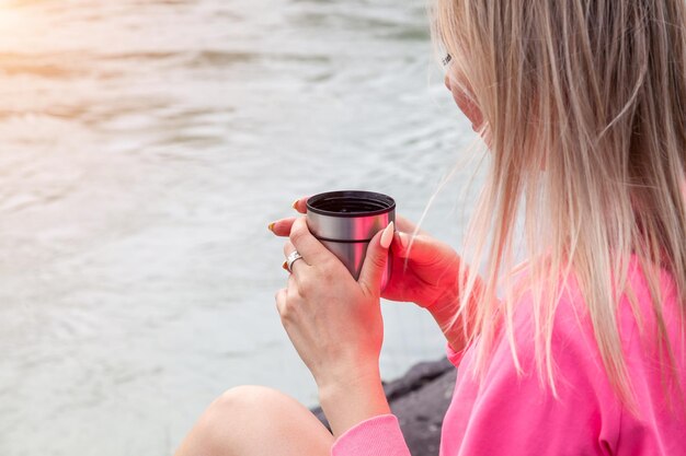 A young beautiful blonde girl in a pink suit with shorts sits on a large stone near the river in the altai mountains and drinks hot tea from an iron mug while relaxing