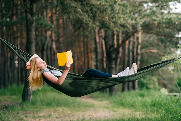 Young beautiful blonde girl reading book in hammock at nature