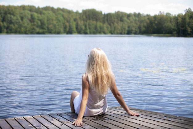 Молодая красивая женщина со светлыми волосами сидит на деревянном пирсе и смотрит на озеро