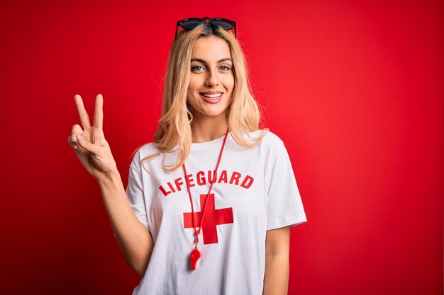 Photo young beautiful blonde lifeguard woman wearing tshirt with red cross using whistle smiling with happy face winking at the camera doing victory sign number two