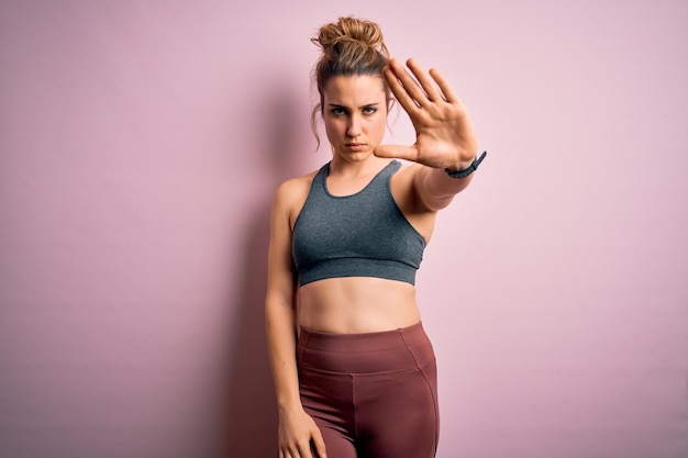 Young beautiful blonde sportswoman doing sport wearing sportswear over pink background doing stop sing with palm of the hand Warning expression with negative and serious gesture on the face