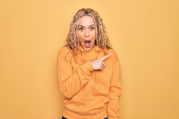 Young beautiful blonde sporty woman wearing casual sweatshirt over yellow background surprised pointing with finger to the side open mouth amazed expression