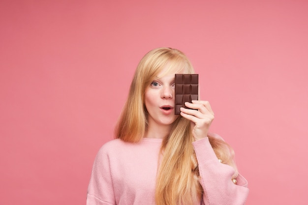 チョコレートと若い美しいブロンド。十代の少女はチョコレートをかみます。禁断のチョコレートを食べる誘惑。チョコレートバーを保持している陽気な肯定的な若い女の子。