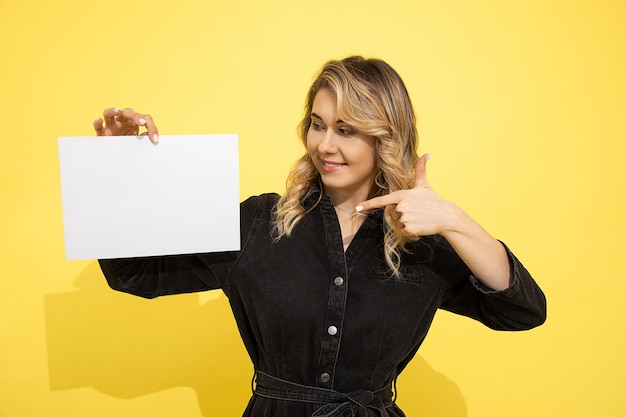 Young beautiful blonde woman in black dress holds empty sheet for inscription on yellow background in hands pointing with her finger, smiles and laughs happily on yellow background in studio