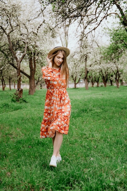 Young beautiful blonde woman in blooming garden. Spring trees in bloom. Orange dress and straw hat.