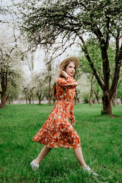 ブルーミングガーデンの若い美しいブロンドの女性。春の木が咲いています。オレンジ色のドレスと麦わら帽子。