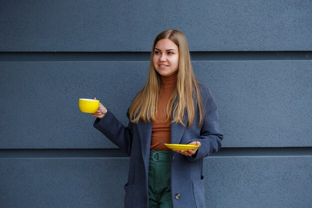 Young beautiful blonde woman in a gray coat drinks hot tea from a yellow cup on a cold autumn day. Delicious warm drink on the terrace in the cafe