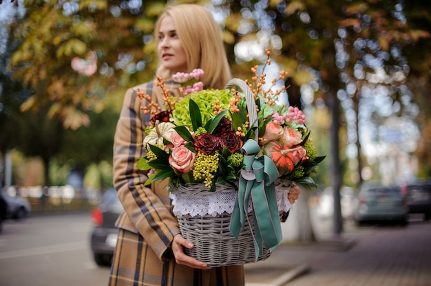 Giovane e bella donna bionda che tiene un grande cesto di vimini di fiori contro la città