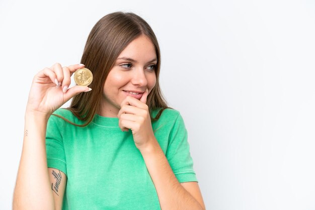 Young beautiful blonde woman holding a Bitcoin isolated on white background thinking an idea and looking side